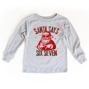 May include: A light gray long-sleeved shirt featuring a graphic of Santa Claus wearing sunglasses and making a peace sign. The text "SANTA SAYS" and "SIX SEVEN" are printed in red and green.
