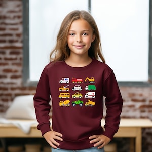 Pode incluir: Uma camisola de cor vinho com um estampado gráfico de vários veículos, incluindo um autocarro escolar, um carro da polícia, um camião de bombeiros, um camião de construção e um camião monstro.