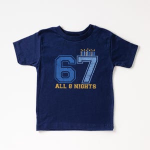 May include: Navy blue toddler t-shirt with the number 67 in blue and gold, topped with lit candles. The text "ALL 8 NIGHTS" is printed below the numbers in gold. The shirt is made of a soft material.