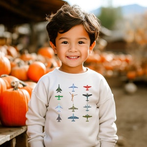 Puede incluir: Un niño pequeño con una sudadera blanca con un estampado colorido de aviones en la parte delantera. El niño sonríe y está de pie frente a una pila de calabazas.