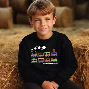 以下が含まれることがあります： カラフルな電車グラフィック、カボチャ、そして「HALLOWEEN EXPRESS！」の文字が特徴の黒いスウェットシャツ。電車の車両は紫、オレンジ、緑など様々な色です。背景には干し草の俵が写っています。