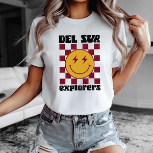 May include: White t-shirt featuring a retro design with the text "DEL SUR explorers". The design includes a yellow smiley face with lightning bolts and a burgundy and white checkered background.