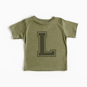 Puede incluir: Camiseta verde oliva para niño con la letra "L" en verde oscuro, de estilo universitario. La camiseta de manga corta está hecha de tela suave y se muestra sobre un fondo blanco. La letra está centrada en la parte delantera de la prenda.