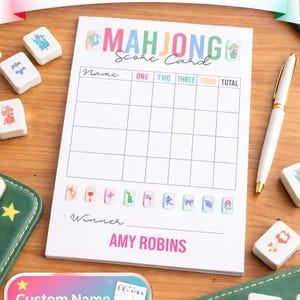 May include: A personalized Mahjong score card with the text "MAHJONG Score Card" and a grid for tracking scores. The card is on a wooden surface, surrounded by Mahjong tiles and a pen. The text "Custom Name AVAILABLE" is also visible.