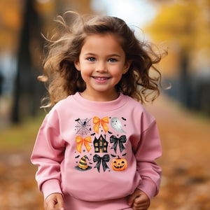 Op de afbeelding: Een jong meisje draagt een roze sweatshirt met een Halloween-thema. Het ontwerp omvat een spinnenweb, oranje strikken, een spook, een spookhuis, zwarte strikken, een heksenhoed en een pompoen.