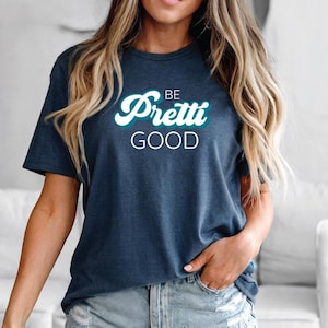 May include: Navy blue t-shirt with the words "BE Pretti GOOD" in a white and teal font. The shirt is a casual style, suitable for everyday wear.