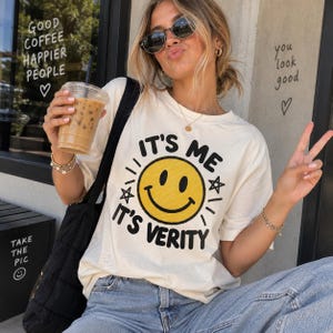 Könnte beinhalten: Ein cremefarbenes Kurzarm-T-Shirt mit einer großen gelben Smiley-Grafik in der Mitte. Der Text "IT'S ME IT'S VERITY" ist in schwarzen Großbuchstaben über und unter dem Smiley aufgedruckt, ergänzt durch kleine schwarze Sternsymbole.
