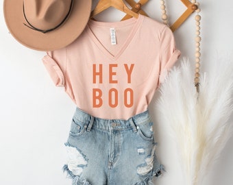 May include: A peach colored v-neck t-shirt with the text "HEY BOO" in orange. The shirt is hanging on a wooden hanger and is paired with denim shorts.