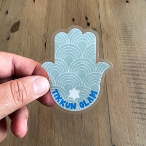 May include: A clear sticker with a blue hamsa hand design. The hamsa hand has a white star in the center and the words "Tikkun Olam" written in blue around the star.