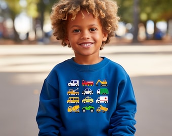 May include: A young boy wearing a blue sweatshirt with a colorful pattern of various vehicles on the front. The vehicles include a school bus, a police car, a fire truck, a tow truck, a construction truck, and a delivery truck.