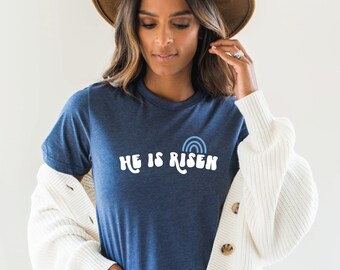 Könnte beinhalten: Ein marineblaues T-Shirt mit einer weißen Grafik, auf der "He is Risen" steht, mit einem blauen Regenbogen über dem Text.