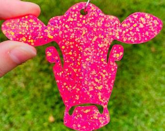 Könnte beinhalten: Ein hellrosa kuhkopf-förmiger Schlüsselanhänger mit Glitzer. Der Schlüsselanhänger wird von einer Hand vor einem grünen Gras Hintergrund gehalten. Der Kuhkopf hat oben ein Loch zum Befestigen an einem Schlüsselring.