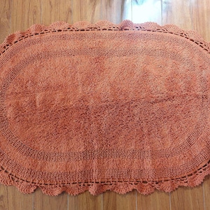 May include: Oval-shaped, coral-colored bath mat with a textured surface and a decorative, scalloped crochet border. The rug is placed on a wooden floor, showcasing its design and color.