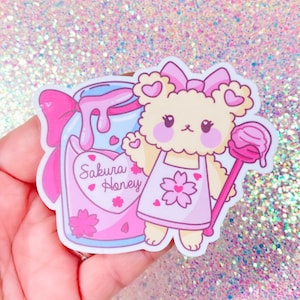 May include: A pink and white sticker featuring a cartoon bear wearing a pink apron with a cherry blossom design. The bear is holding a pink and white lollipop and a jar of pink liquid with the text "Sakura Honey".