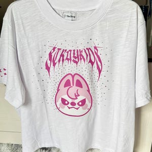 Puede incluir: Camiseta blanca con un gráfico rosa que presenta el texto "Stray Kids" en una fuente estilizada y una cara de personaje de dibujos animados rosa. La camiseta tiene cuello redondo y mangas cortas. La marca "The Drop" está impresa en la etiqueta.