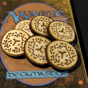 Puede incluir: Seis fichas de madera con un diseño circular, con un recorte central en forma de Y y pequeñas perforaciones. Están dispuestas sobre una carta de Magic: The Gathering, con la palabra "Deckmaster" visible.
