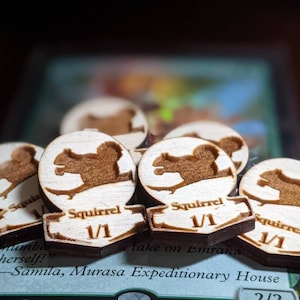 May include: Close-up of five wooden tokens with a squirrel silhouette and the text "Squirrel 1/1" on each. The tokens are arranged in a semi-circle on a green and brown card with the text "take on Emraka" and "Samila, Murasa Expeditionary House".