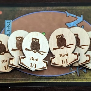 Könnte beinhalten: Helle Vogel-Token aus Holz mit einem gravierten Eulen-Design. Jeder Token zeigt eine Illustration einer sitzenden Eule, das Wort "Bird" und den Text "1/1". Die Token sind aus hellem Holz mit dunkelbraunen Gravuren, geeignet für Tabletop-Spiele.