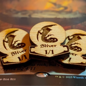 May include: Three wooden tokens with a circular top and a rectangular base. Each token features a carved illustration of a creature and the word "Sliver" above the numbers "1/1". The tokens are made of light-colored wood.