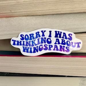 May include: A white sticker with blue text that reads "Sorry I was thinking about wingspans". The sticker is placed on a stack of books.