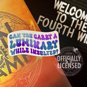 May include: A white sticker with a colorful, wavy font that reads "Can you carry a luminary while insulted?" The sticker is placed on a book with a gold and orange cover. The book has the text "The Empyrean" and "Officially Licensed" on the cover.