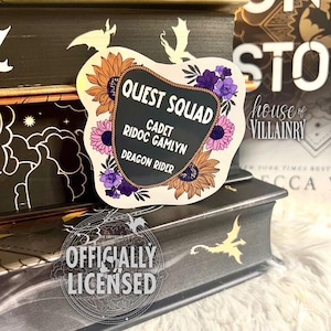 May include: A white sticker with a black chalkboard-style sign that reads "Quest Squad" with the text "Cadet Ridoc Gamlyn Dragon Rider" underneath. The sticker is decorated with sunflowers, purple flowers, and dragon silhouettes.
