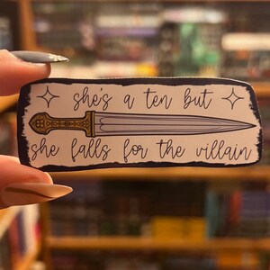 May include: A white sticker with a black border and a gold and silver dagger. The sticker says "She's a ten but she falls for the villain" with a star on each side of the text.