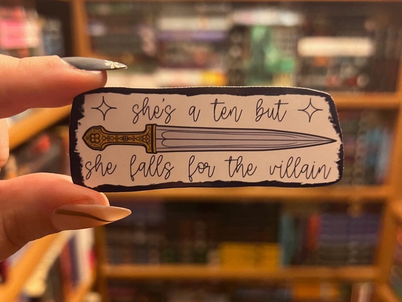 May include: A white sticker with a black border and a gold and silver dagger. The sticker says "She's a ten but she falls for the villain" with a star on each side of the text.