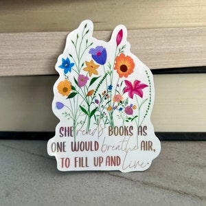 May include: A white sticker featuring a colorful floral design with various flowers in shades of blue, orange, pink, and yellow. The sticker has the quote, "She reads books as one would breathe air, to fill up and live."