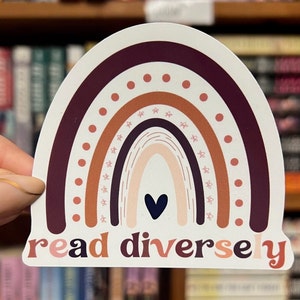 May include: A sticker featuring a rainbow design with the words "read diverse y". The rainbow has layers of burgundy, orange, and tan colors, with a navy blue heart at the bottom. The sticker is held in front of a bookshelf.