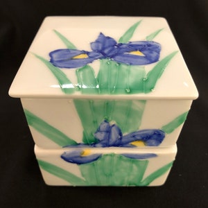 May include: A white ceramic two-tiered box with a floral design. The box features blue and yellow irises with green leaves.