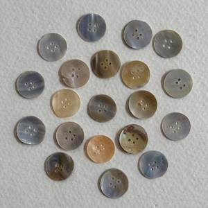 May include: A collection of round, iridescent buttons in various shades of gray, tan, and peach. Each button has four holes for sewing. The buttons are arranged on a light-colored surface.