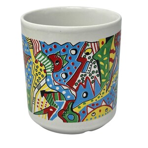 May include: White ceramic mug with a colorful abstract design. The design features blue, red, yellow, and green shapes and patterns. The mug has a cylindrical shape and a smooth, glossy finish. The mug is suitable for hot or cold beverages.