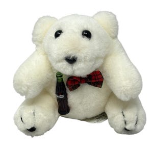 May include: A white plush polar bear toy with black eyes and nose, wearing a red and black plaid bow tie. The bear holds a miniature Coca-Cola bottle. The toy is made of soft, fluffy material.