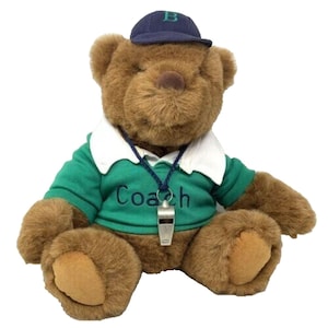May include: A brown teddy bear wearing a green and white polo shirt with the word "Coach" embroidered on it. The bear is also wearing a blue baseball cap with a white letter "B" on it. The bear has a blue lanyard with a silver whistle attached to it.