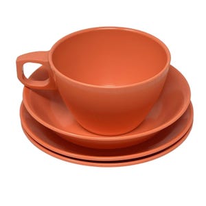 May include: A set of coral-colored ceramic tableware, including a large teacup with a handle and two matching saucers. The cup and saucers are stacked, showcasing a simple, modern design. The set is suitable for serving tea or coffee.