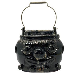 May include: A black ceramic container with a face design, featuring large round eyes, a nose, and a smiling mouth. It has a metal handle and small round accents. The container is likely for decorative or functional use.