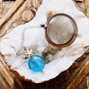 May include: A silver tea infuser with a chain and a blue glass bead with a white starfish charm. The infuser is sitting in a white crystal geode on a wooden surface.