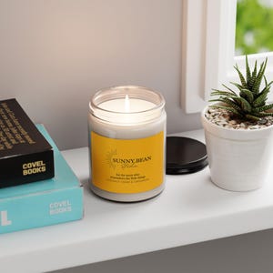 May include: A lit candle in a clear glass jar with a yellow label that reads "SUNNY BEAN Studio" and "COCONUT CREAM & CARDAMOM". The candle sits on a white shelf next to a small potted succulent and two books.