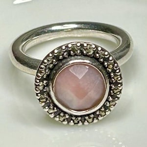 May include: A silver ring featuring a round, faceted, pale pink stone at its center. The stone is surrounded by a circular border of small, dark, metallic-toned stones set within a silver frame, attached to a smooth, polished silver band.