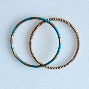 Op de afbeelding: Twee gouden armbanden, een met een turquoiseblauw emaille ontwerp en de andere met een textuur van goud.
