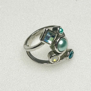 Könnte beinhalten: Ein silberner Ring mit einzigartigem Design, mit einem Quadrat, einem Kreis und zwei ovalen Fassungen, die jeweils einen andersfarbigen Edelstein halten. Die Edelsteine umfassen hellblau, türkis und irisierendes Weiß. Der Ring ist mit "lie soph" markiert.