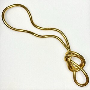 Peut inclure: Un collier doré avec un design noué. Le collier est composé d'une chaîne souple en forme de serpent. La chaîne est d'une couleur or brillante et brillante. Le collier est présenté sur un fond blanc.