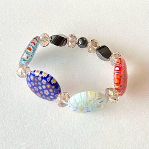 May include: A colourful bracelet with round glass beads featuring floral patterns in blue, red, and white. The bracelet also includes clear and black beads.