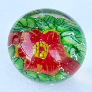 May include: A clear glass paperweight featuring a red and yellow flower design. The flower is surrounded by green leaves, and small air bubbles are visible. The paperweight is round in shape, with a diameter of approximately 7 cm.