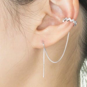 Peut inclure: Un ensemble de boucles d'oreilles et de manchette d'oreille argentées. La manchette d'oreille a un design en zigzag et se place sur le haut de l'oreille. Une chaîne relie la manchette à une boucle d'oreille pendante avec une barre droite.