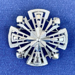 Puede incluir: Un broche plateado en forma de cruz con un diseño geométrico. La cruz tiene un patrón de estrella en el centro.