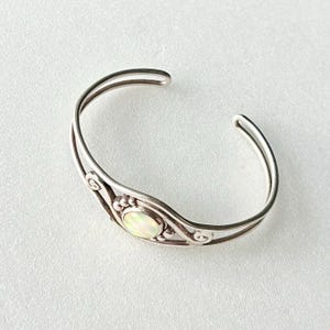 Peut inclure: Bracelet manchette en argent avec une pierre précieuse ovale et irisée au centre. Le bracelet présente un design délicat et ouvert avec des volutes décoratives et une finition polie. Un bijou classique.