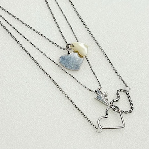 May include: A layered necklace with three strands. The top strand features a heart pendant and a shell-shaped charm. The middle strand has an arrow pendant with a small gem. The bottom strand has a heart-shaped pendant.