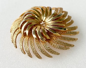 May include: A gold-toned brooch with a swirling, sunburst design. The brooch is made of metal and has a textured finish.